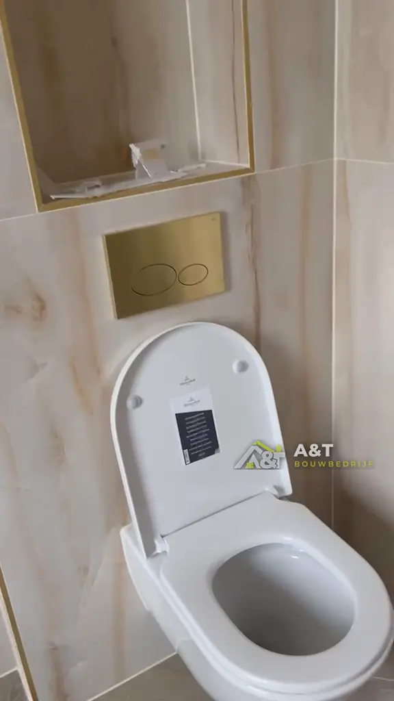 Inbouwtoilet met gouden plaat. Marmerlook-tegels, nis met gouden profiel en hangtoilet.